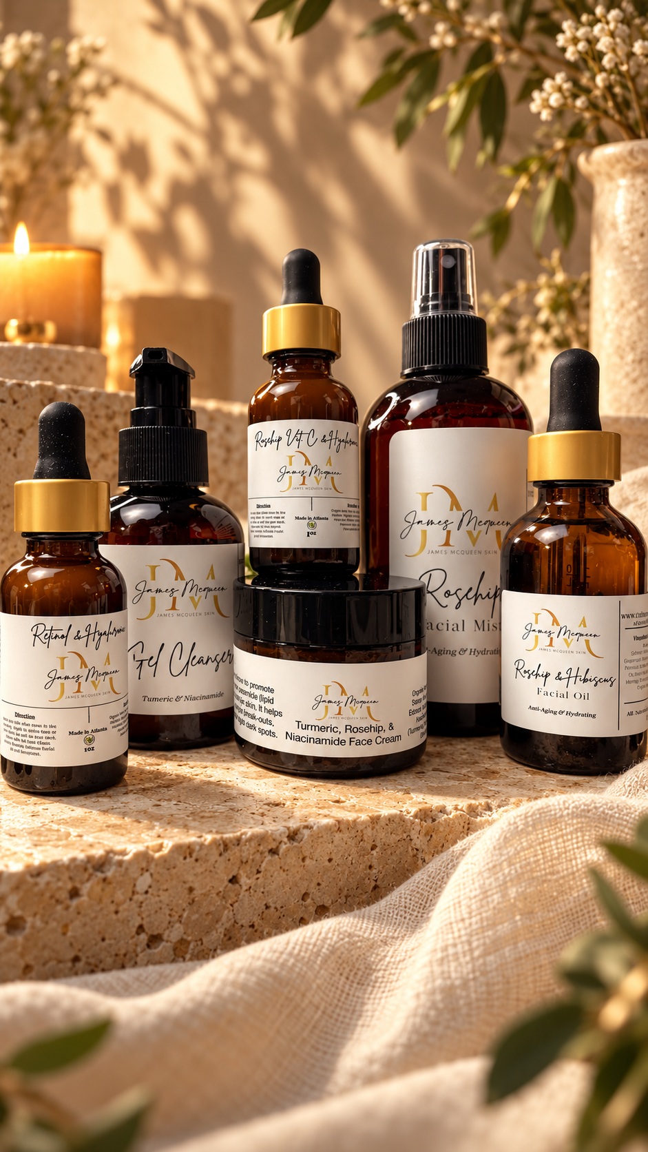 Six skincare bottles and jars from the brand James McQueen Skin arranged on a stone ledge with a linen cloth and candles in the background.